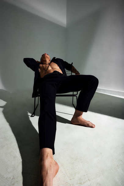 Weitwinkelporträt eines jungen brünetten Mannes in schwarzem Hemd und schwarzer Hose, mit muskulösem Oberkörper, der entspannt auf einem Stuhl sitzt und aufblickt. Stilvolles Studioshooting - Foto, Bild