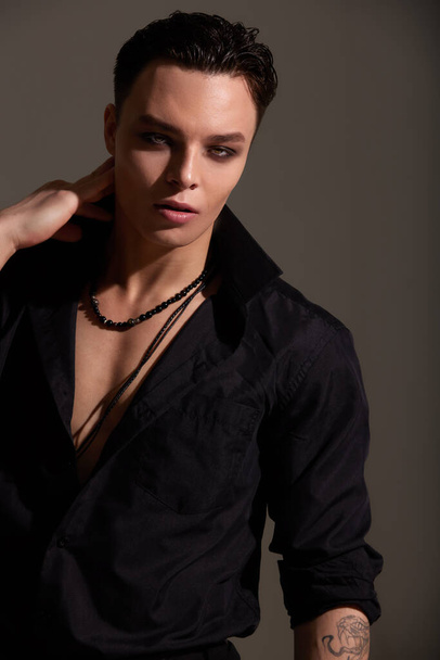 Studio portrait of a young teenager brunette boy in a black shirt with beads around his neck. Short trendy haircut. Elegant sexy style. Gray background - Foto, afbeelding