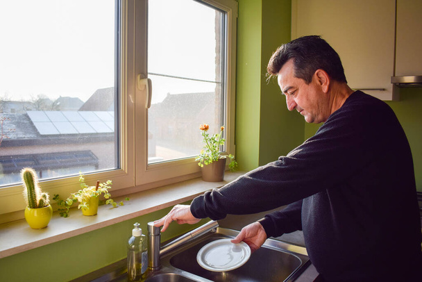 Καυκάσιος ώριμος άντρας πλένει πιάτα στην κουζίνα στο σπίτι μετά το μεσημεριανό. Ο σύζυγος βοηθάει τη σύζυγο να καθαρίσει το σπίτι. Οικιακός καθαρισμός εσωτερικών χώρων. Άνθρωποι που ζουν μόνοι, καθαρίστριες. - Φωτογραφία, εικόνα