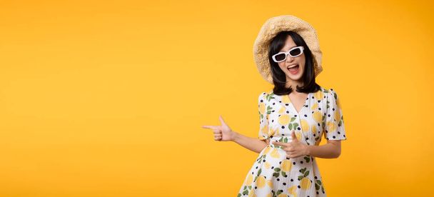 Portrait young beautiful asian woman wearing springtime dress and pointing to copy space for advertising isolated on yellow background. Summertime advertising banner concept. - Foto, Imagen