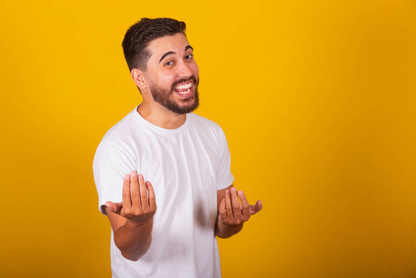 Brazilian Latin American man, with gesture calling, come, come, inviting, receiving, welcoming, happy, smiling, autruism, yellow background, space for text or advertisement - Foto, imagen