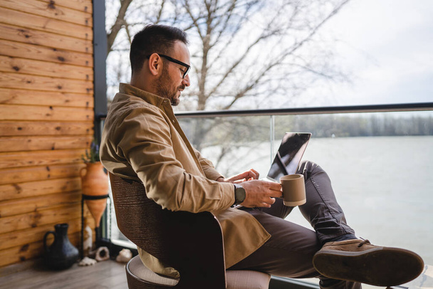 egy férfi felnőtt férfi 40 éves ülni egy teraszon erkély egy csésze kávé használata laptop számítógép vállalkozó vagy szabadúszó nyaralás reggel rutin valós személy másolás hely - Fotó, kép
