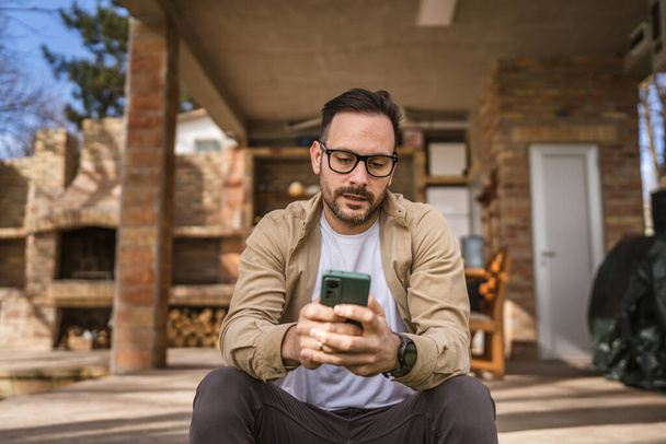 Yksi aikuinen mies 40-vuotias valkoihoinen mies istuu talonsa edessä käyttää älypuhelinta lukemaan viestejä tai uutisia syvällä ajatuksissa huolissaan tai huolissaan ajattelemalla todellisen henkilön kopiotilaa. - Valokuva, kuva