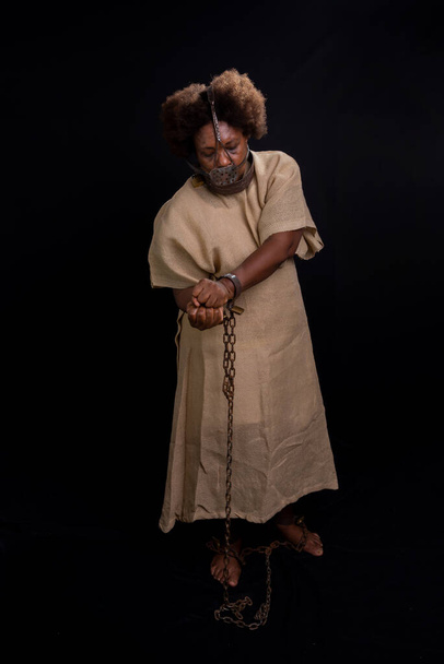 Mujer negra, de pie, encadenada y con una máscara de hierro en la cara que representa a la esclava Anastacia. La esclavitud en Brasil. Aislado sobre fondo negro. - Foto, imagen