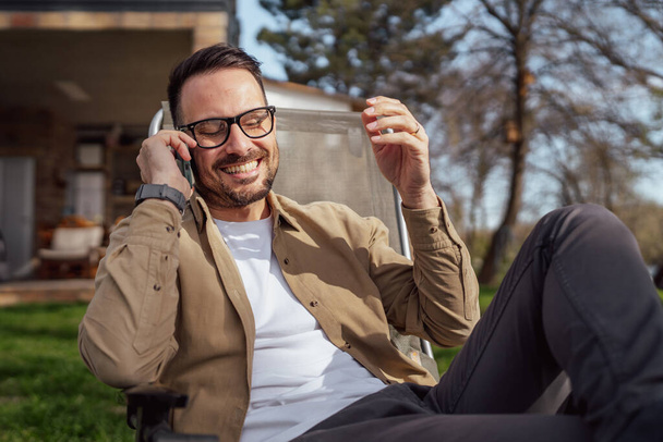 Een volwassen man 40 jaar oud blanke man zitten voor zijn huis op vakantie praten op zijn smartphone mobiele telefoon bellen vrolijke glimlach tijdens het gesprek echte persoon kopiëren ruimte - Foto, afbeelding