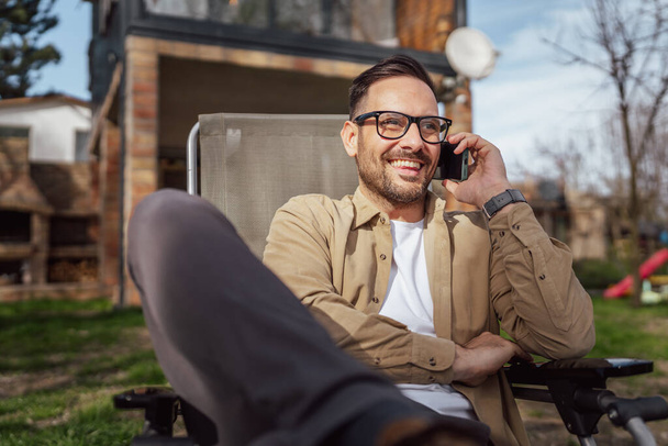 Один дорослий чоловік 40 років білий чоловік сидить перед своїм будинком у відпустці розмовляє на своєму смартфоні мобільний телефонний дзвінок щаслива посмішка під час розмови реальна людина копіює простір
 - Фото, зображення