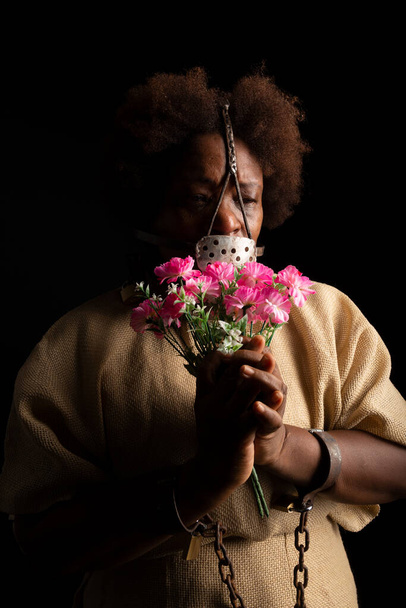 Ellerinde zincirler ve yüzünde bir demet çiçek tutan demir bir maske olan siyah bir kadının portresi. Köle Anastacia. Brezilya 'da kölelik. - Fotoğraf, Görsel