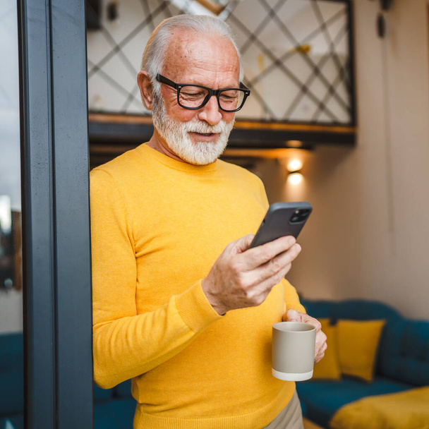 Jeden starszy człowiek ze stojakiem na brodę na balkonie trzymać telefon komórkowy i filiżankę kawy codziennie rano rutyna prawdziwa osoba kopiować przestrzeń czytać lub używać smartfona Internet przeglądać sieci społecznościowe - Zdjęcie, obraz