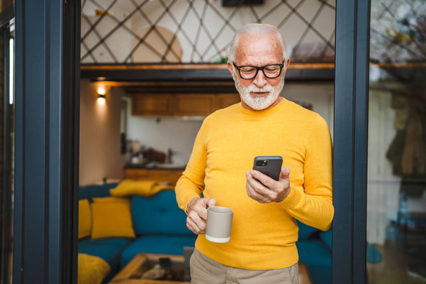 One senior man with a beard stand on the balcony hold a mobile phone and cup of coffee daily morning routine real person copy space read or use smartphone internet browse social network - Foto, imagen
