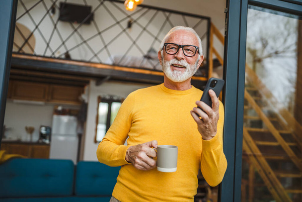 Un homme âgé avec un stand de barbe sur le balcon tenir un téléphone mobile et une tasse de café sourire heureux tous les jours matin routine personne réelle copier l'espace lu ou utiliser smartphone Internet naviguer sur le réseau social - Photo, image