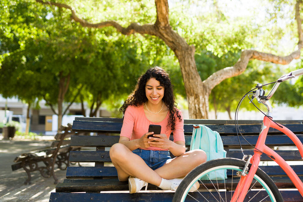Pihent gyönyörű fiatal nő ül a parkban padon, és sms-ezik az okostelefonon, miután lovagolt a kerékpár a szabadban - Fotó, kép