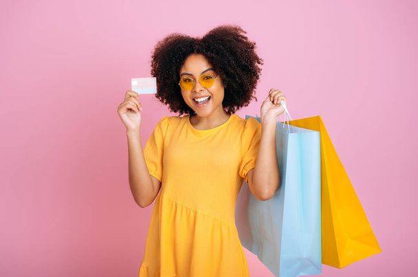 Aufgeregt fröhliche brasilianische oder afrikanisch-amerikanische lockige Frau, in einem gelben Sommerkleid, Papiertüten mit Einkäufen und einer Kreditkarte in der Hand, erstaunten Blicken in die Kamera, Lächeln, stehen auf isoliertem rosa Hintergrund - Foto, Bild