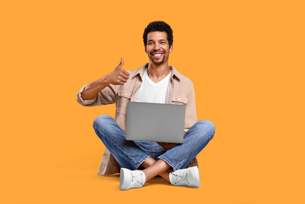 Happy man with laptop showing thumb up against orange background - Valokuva, kuva