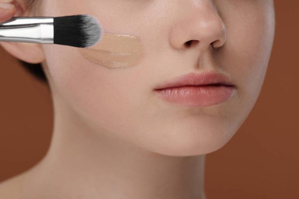 Teenage girl applying foundation on face with brush against brown background, closeup - Foto, imagen