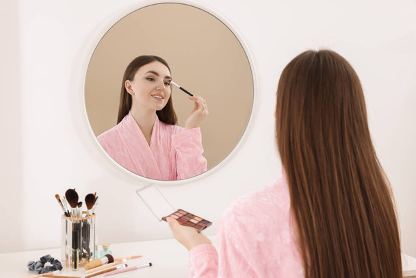 Beautiful young woman applying eyeshadow with brush near mirror indoors - Valokuva, kuva