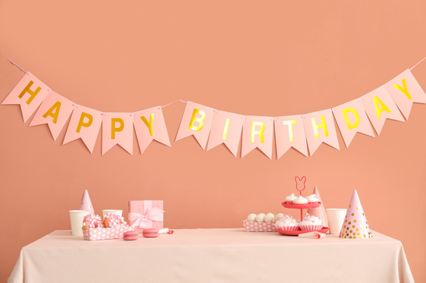 Édességek fehér asztalon és boldog születésnapot papír koszorúk sötét bézs falon - Fotó, kép