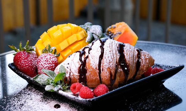 heerlijke Franse ontbijt croissant met chocolade en poedersuiker op een zwarte plaat in een restaurant op straat Mango fruit bessen frambozen aardbeien papaja munt mooie portie ochtend - Foto, afbeelding