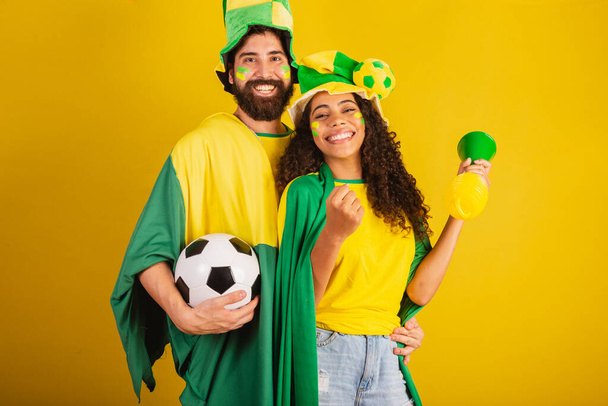 Пара бразильских футбольных болельщиков, одетых в цвета нации, черная женщина, белый мужчина. использование флага и аксессуаров - Фото, изображение