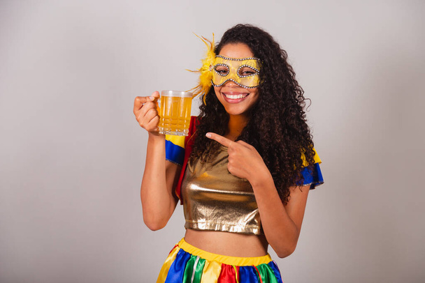 Beautiful black Brazilian woman, with frevo clothes, carnival. in mascara, showing glass of beer, partying in bar. - Fotoğraf, Görsel