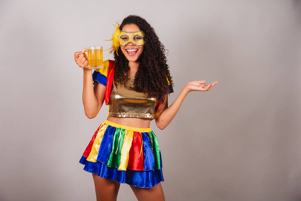 Beautiful black Brazilian woman, with frevo clothes, carnival. wearing a mask, dancing with a glass of beer, partying in a bar. - Foto, imagen