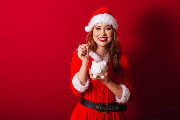 beautiful Brazilian red-haired woman, dressed in Christmas clothes, Santa Claus. holding piggy bank and coin. - Foto, afbeelding