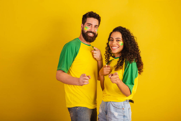 coppia di tifosi brasiliani, vestiti con i colori della nazione, donna nera, uomo caucasico. Indicando la fotocamera. - Foto, immagini