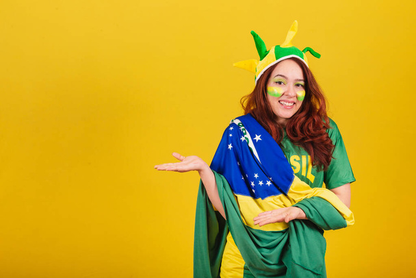 Caucasian, red-haired woman, soccer fan from Brazil, presenting products with her hands. - Zdjęcie, obraz