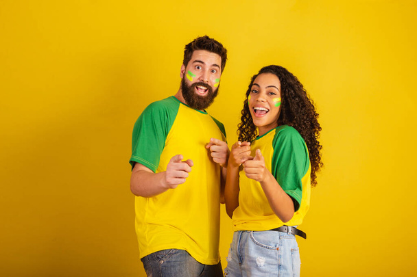 couple of brazil soccer supporters, dressed in the colors of the nation, black woman, caucasian man. Pointing at camera. - Foto, immagini