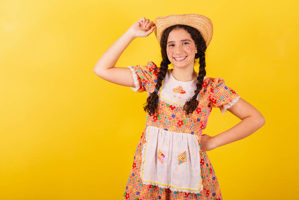 Girl wearing traditional orange clothes for festa junina. Smiling with hand in hat, greeting. - 写真・画像