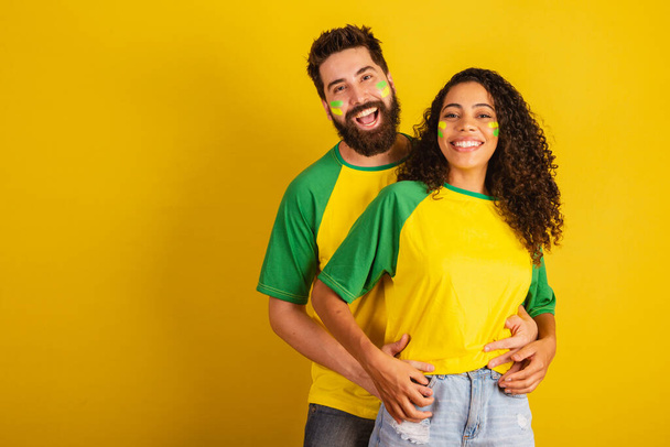 couple of brazil soccer supporters, dressed in the colors of the nation, black woman, caucasian man. embraced. - Foto, afbeelding