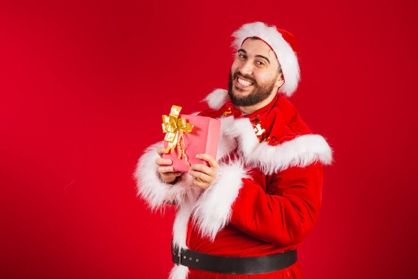 brazilian man, dressed in santa claus clothes, holding red gift. - Foto, Bild