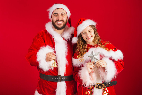 Brazilian couple, dressed in Christmas clothes, Santa Claus, holding Christmas decorations, star, bell. - Foto, imagen