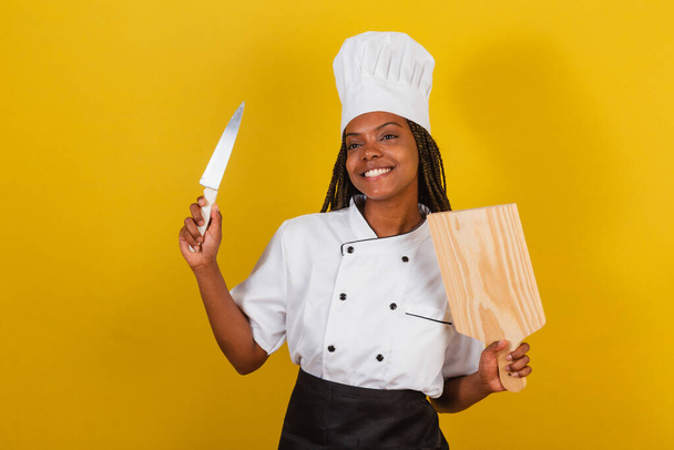 Young afro brazilian woman, chef cook, holding wooden board and knife. - 写真・画像
