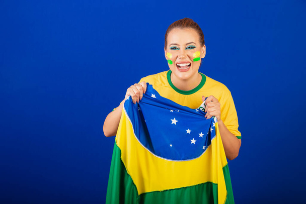 Caucasian woman, redhead, Brazil soccer fan, Brazilian, blue background, screaming goal with flag in hands - Photo, Image