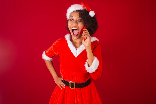 beautiful black brazilian woman, dressed as santa claus, mama claus, talking on voice call by smartphone, surprised, wow, amazing - Foto, imagen