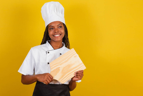 Young afro brazilian woman, cook, holding wooden board for gastronomy. - 写真・画像