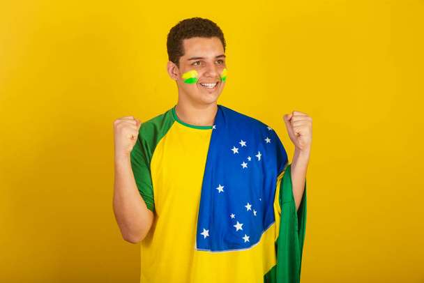 Young Brazilian man soccer fan. dressed in green, hand on chest, celebrating with the flag of Brazil. - Foto, imagen