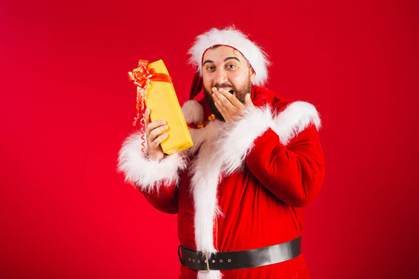 brazilian man, dressed in santa claus clothes, doubting what's in yellow gift, doubt, surprise. - Zdjęcie, obraz