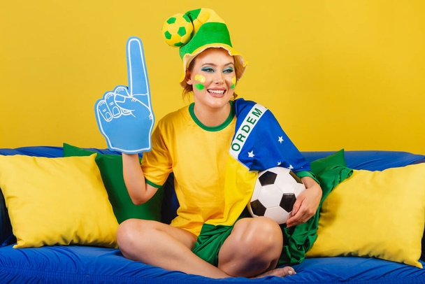 Caucasian, redhead, Brazilian, Brazilian soccer fan on couch with foam finger and ball partying - Foto, afbeelding
