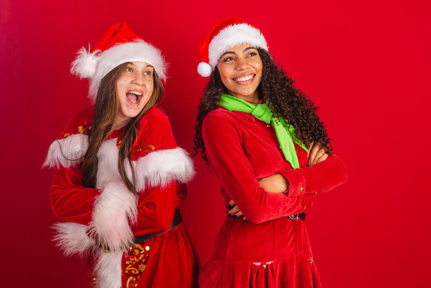 İki Brezilyalı bayan arkadaş, Noel Baba kıyafetleri giymişler, kolları iyimser bir şekilde çaprazlanmış gülümsüyorlar.. - Fotoğraf, Görsel