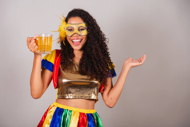 Beautiful black Brazilian woman, with frevo clothes, carnival. wearing a mask, dancing with a glass of beer, partying in a bar. - Foto, immagini