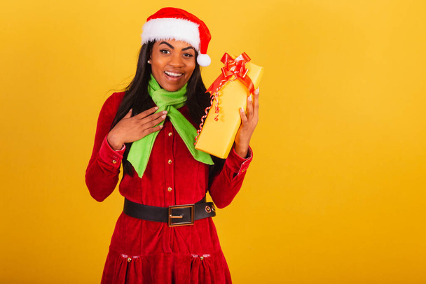 Beautiful black Brazilian woman, dressed in Christmas clothes, Santa Claus, hugging yellow gift. - Zdjęcie, obraz