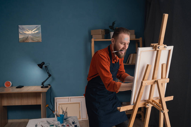 portrait caucasian bearded male artist in an apron in a home studio stands near an easel holds a canvas in his hands and looks at the picture contemptuously. hobby and vacation concept. - Zdjęcie, obraz