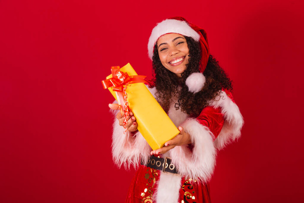 beautiful brazilian black woman, dressed as santa claus, christmas clothes. gifting someone with yellow gift. - Zdjęcie, obraz