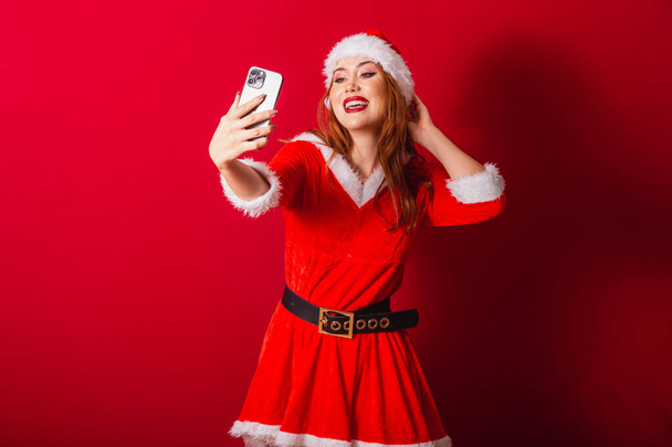 beautiful Brazilian red-haired woman, dressed in Christmas clothes, Santa Claus. taking self portrait through smartphone. - Φωτογραφία, εικόνα