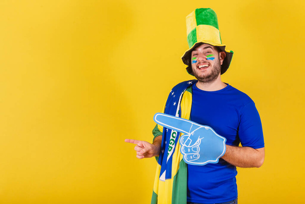 Brazilian caucasian man, soccer fan from Brazil, wearing hat, using foam finger to point left. - Foto, imagen