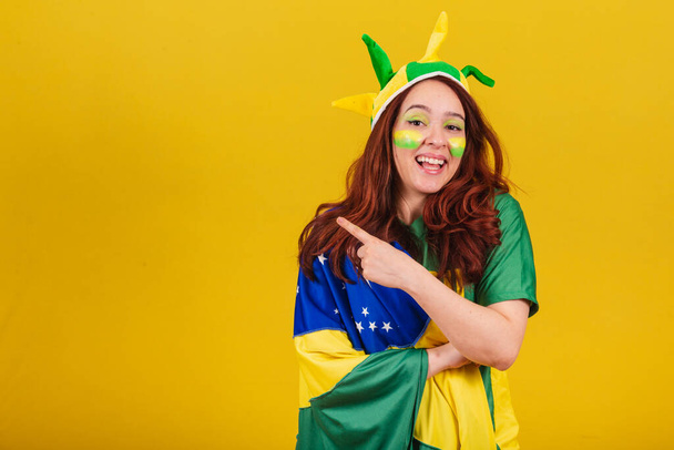 Caucasian, red-haired woman, soccer fan from Brazil, pointing with index finger to the left. Advertising photo. - Foto, Bild