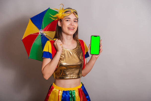 Beautiful Brazilian, Caucasian woman, wearing frevo, carnival clothes, wearing a mask. holding smartphone with green screen in chroma. - Photo, Image