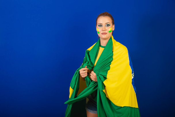 Mujer caucásica, pelirroja, aficionada al fútbol de Brasil, brasileña, fondo azul, usando la bandera como capa - Foto, imagen