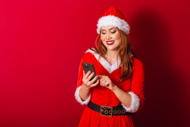 beautiful Brazilian red-haired woman, dressed in Christmas clothes, Santa Claus. holding smartphone, typing. - Φωτογραφία, εικόνα